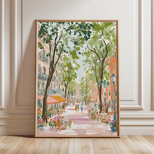 May include: A framed watercolour painting of a European street scene. The artwork shows a tree-lined boulevard with cafes, people, and buildings in shades of pink, orange, and green. The painting is in a wooden frame.
