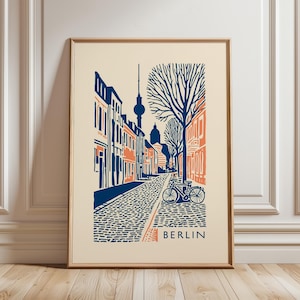 May include: Framed art print depicting a Berlin street scene in a stylized illustration. The artwork features buildings in blue and orange, a cobbled street, a bicycle, and the TV Tower. The word "BERLIN" is printed at the bottom of the print.