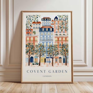Op de afbeelding: Een ingelijste aquarelprint van Covent Garden, Londen. Het kunstwerk toont kleurrijke gebouwen met oranje, blauwe en witte gevels en bomen met sinaasappels. De tekst "COVENT GARDEN LONDON" staat onderaan.