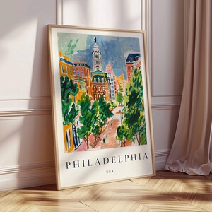 May include: A framed art print with a watercolour painting of a Philadelphia street scene. The artwork shows buildings, trees, and a tower, with the city name and "USA" printed at the bottom. The frame is light-coloured.
