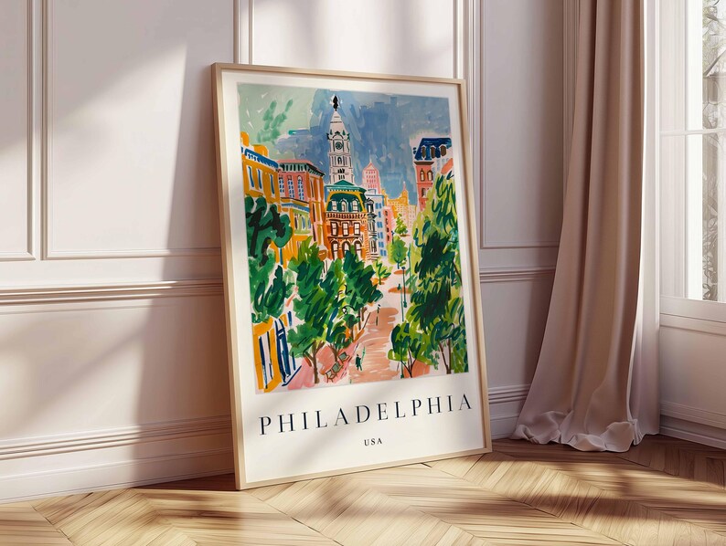 May include: A framed art print with a watercolour painting of a Philadelphia street scene. The artwork shows buildings, trees, and a tower, with the city name and "USA" printed at the bottom. The frame is light-coloured.