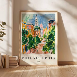 May include: A framed art print showing a vibrant illustration of Philadelphia, USA. The artwork depicts a street scene with buildings, trees, and a clock tower. The word "PHILADELPHIA" is printed at the bottom.