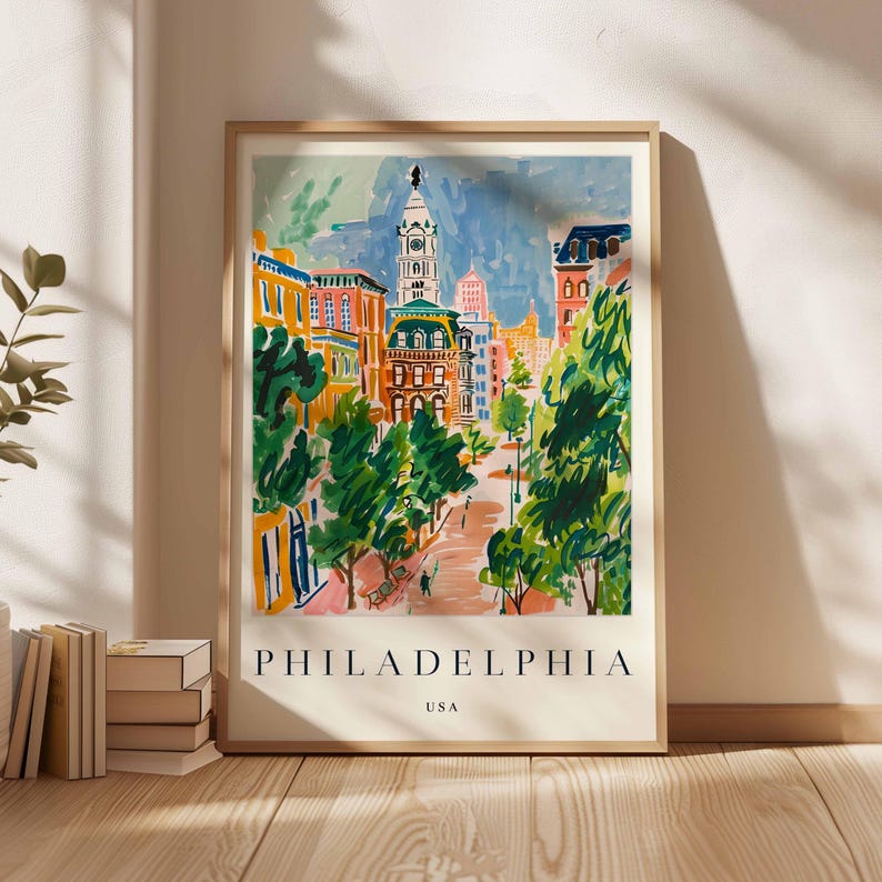 May include: A framed art print showing a vibrant illustration of Philadelphia, USA. The artwork depicts a street scene with buildings, trees, and a clock tower. The word "PHILADELPHIA" is printed at the bottom.