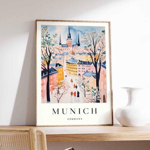 Puede incluir: Una ilustración de acuarela de una ciudad con edificios de colores pastel y un campanario de iglesia. La ilustración está enmarcada en un marco de madera clara y tiene el texto "MUNICH GERMANY" en la parte inferior.