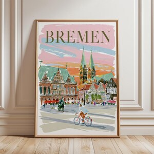 May include: A framed art print featuring a painterly illustration of Bremen, Germany. The scene depicts a city square with historic buildings, tall spires, and a person riding a red bicycle. The sky is painted in soft pink and blue brushstrokes.