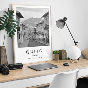 May include: A black and white photograph of a street in Quito, Ecuador. The photo is framed in a light wood frame and is displayed on a desk with a laptop, a lamp, and a potted plant. The text "QUITO" and "ECUADOR" is printed below the photo, along with the coordinates "0.1807&deg; S, 78.4678&deg; W".