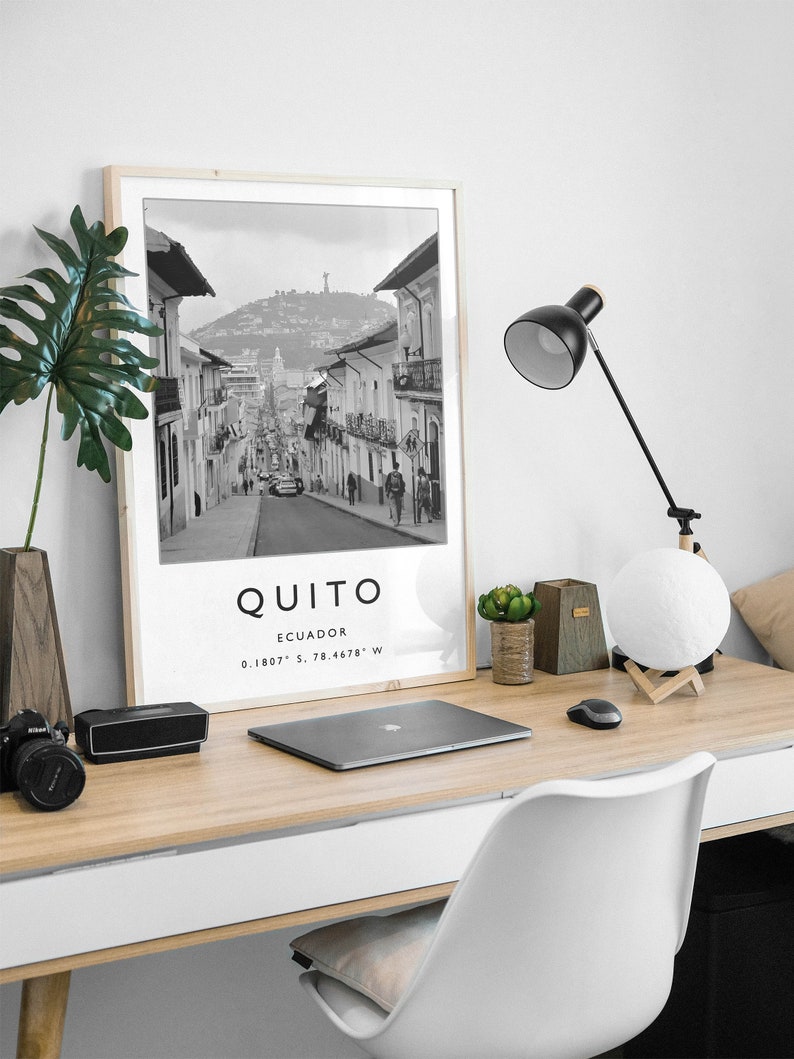 May include: A black and white photograph of a street in Quito, Ecuador. The photo is framed in a light wood frame and is displayed on a desk with a laptop, a lamp, and a potted plant. The text "QUITO" and "ECUADOR" is printed below the photo, along with the coordinates "0.1807&deg; S, 78.4678&deg; W".