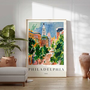 May include: A framed watercolour print of Philadelphia, USA, depicting a colourful cityscape with buildings, trees, and a blue sky. The print is displayed on a wooden floor, with a potted plant and decorative vases nearby.