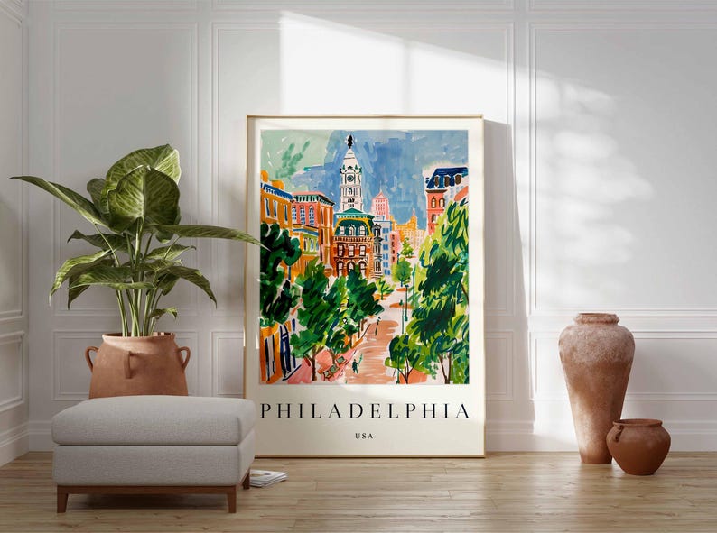 May include: A framed watercolour print of Philadelphia, USA, depicting a colourful cityscape with buildings, trees, and a blue sky. The print is displayed on a wooden floor, with a potted plant and decorative vases nearby.