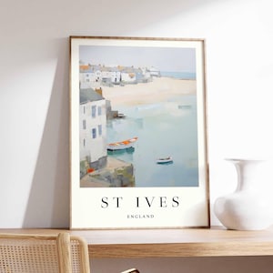 May include: A watercolor painting of a coastal town with white buildings and a blue body of water. The painting is framed in a light wood frame and has the text "ST IVES ENGLAND" at the bottom.