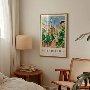 May include: A framed art print of Philadelphia, USA, in a colourful, impressionistic style. The artwork depicts a street scene with buildings, trees, and the city's name at the bottom. The print is displayed on a wall in a room with a lamp and chair.