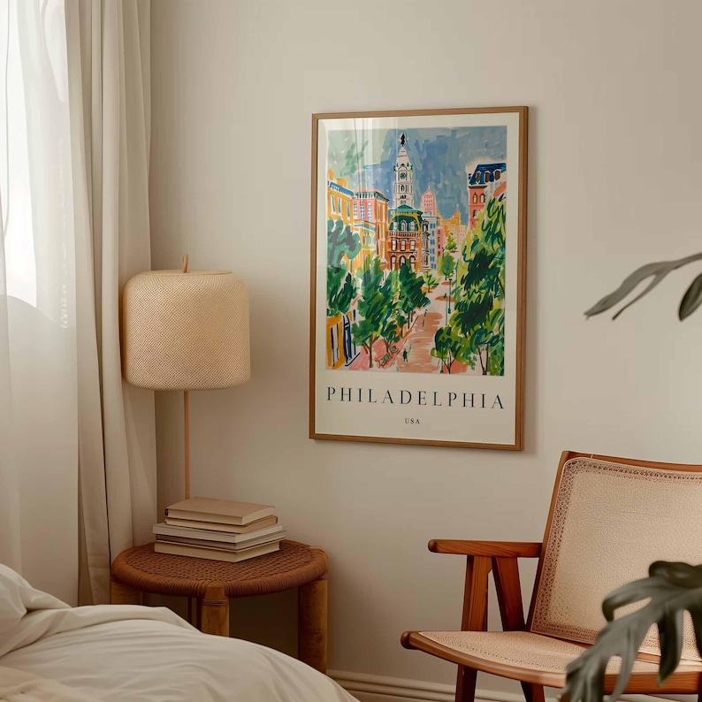 May include: A framed art print of Philadelphia, USA, in a colourful, impressionistic style. The artwork depicts a street scene with buildings, trees, and the city's name at the bottom. The print is displayed on a wall in a room with a lamp and chair.