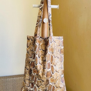 Puede incluir: Un bolso tote rectangular con un patrón de piel de serpiente en tonos marrón, beige y blanco. El bolso cuenta con dos asas de tela a juego y un diseño plano con la parte superior abierta. La superficie muestra un estampado de escamas.