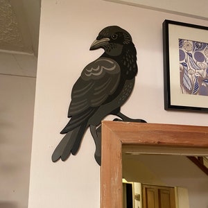 May include: A black crow with a grey and white pattern on its wings is mounted on a white wall. The crow is facing the right side of the image. The crow is made of wood and has a three-dimensional appearance.