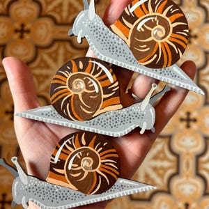 May include: Three flat, hand-painted wooden snail cutouts featuring light grey bodies with dark grey stippled texture and spiralled shells in shades of brown, orange, and cream. Each snail has two prominent tentacles and a distinct, curved shell design.