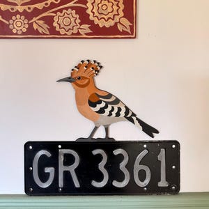 May include: A decorative wall hanging featuring a cutout of a hoopoe bird with orange, black, and white plumage, perched atop a rectangular black metal license plate with the embossed silver-coloured alphanumeric text GR 3361.
