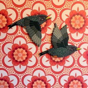 May include: Two wooden bird wall art pieces in flight, with black and white speckled bodies, grey wings, and yellow beaks. The birds are set against a retro floral patterned background in red, orange, and white. Decorative wall art for the home.