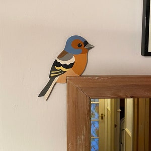 May include: A wooden framed mirror with a blue, orange and black bird cutout on the top. The mirror is reflecting a room with a white door and a wooden floor.