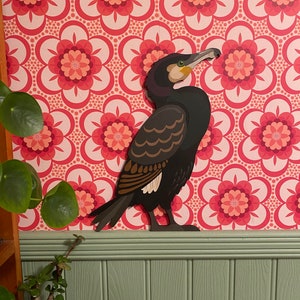 May include: A black and brown paper cut-out of a cormorant bird against a pink and white floral patterned background.