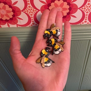 May include: Three wooden bumblebee ornaments with black and yellow stripes and white accents. The bees are arranged in a hand, with the wings of the bees facing outwards.