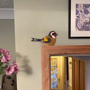May include: A wooden bird figurine with a red head, yellow breast, and black wings. The bird is perched on a wooden shelf above a doorway.