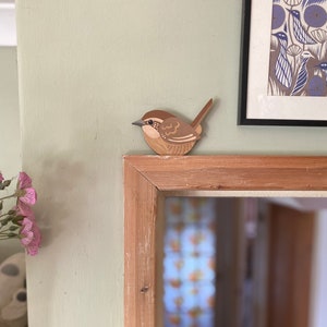 May include: A wooden bird figurine of a wren, painted in shades of brown and beige, perched on a wooden shelf. The bird has a long tail and a small beak.