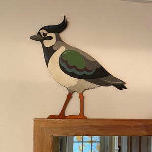 May include: A painted wooden bird decoration. The bird has a distinctive crest, a white chest, and a green and blue wing pattern. The legs are orange, and it is mounted on a wooden surface. The bird is a decorative item.