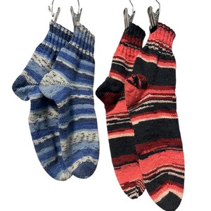 Könnte beinhalten: Zwei Paar gestrickte Socken hängen an einer Holzstange. Ein Paar hat blaue und weiße Streifen, während das andere rote, schwarze und weiße Streifen aufweist. Die Socken werden von Metallklammern gehalten.