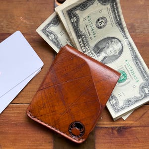 May include: A brown leather wallet with a textured, scratched surface and a small circular metal emblem featuring a crab design. The wallet is shown with two white blank cards and several two-dollar bills tucked behind it.