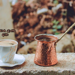 Peut inclure: Une tasse à café blanche avec des détails dorés et une soucoupe, à côté d'une cafetière turque en cuivre avec une longue poignée. L'image comprend le texte "Turkish Coffee Gift". L'arrière-plan est flou.