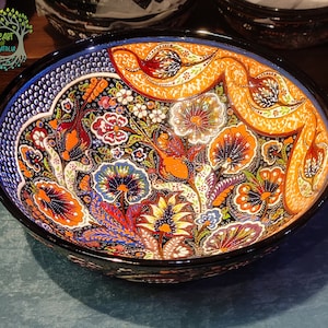 May include: A decorative ceramic bowl with a black rim and a vibrant floral pattern. The bowl features a mix of orange, blue, red, and white flowers and leaves on a black background. The words "Heart of Anatolia" are in the upper left corner.