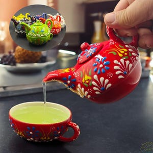 Op de afbeelding: Een rode keramische theepot met een bloemmotief schenkt thee in een bijpassende theekop. De theepot en de theekop hebben witte, blauwe en gele accenten. Een kleine inzet toont andere theepotten.