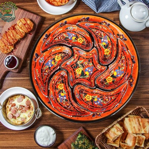 Può includere: Un vivace piatto da portata in ceramica arancione con bordo nero, caratterizzato da un elaborato motivo floreale giallo, blu e bianco. Il piatto è pieno di vari piatti, tra cui patate fritte, uova e pane, disposti su un tavolo di legno.