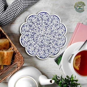 Puede incluir: Un posavasos de cerámica blanca con un patrón floral azul. El posavasos tiene un borde festoneado y es perfecto para proteger las superficies de los platos calientes.