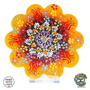 Puede incluir: Un plato decorativo de cerámica con borde festoneado y un vibrante diseño floral. El plato presenta un patrón de mandala central en morado, blanco, verde y naranja, rodeado por un borde amarillo. El texto "MADE WITH LOVE" es visible.