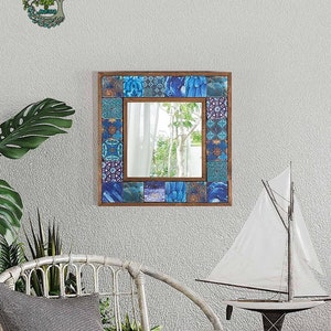 Może przedstawiać: Kwadratowe lustro w drewnianej ramie z obramowaniem z niebieskich i turkusowych płytek mozaikowych. Lustro wisi na teksturowanej szarej ścianie. Widoczne są również białe krzesło z wikliny, model żaglówki i dekoracja ścienna w kształcie drzewa.