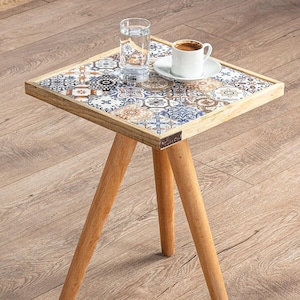 Peut inclure: Une petite table d'appoint en bois avec trois pieds. Le plateau de la table est décoré d'un motif de mosaïque de carreaux colorés. Un verre d'eau et une tasse de café sont sur la table.