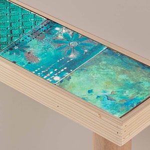 Puede incluir: Una mesa auxiliar de madera con una tapa de patrón turquesa y verde. La mesa tiene una sola pata y un diseño simple.