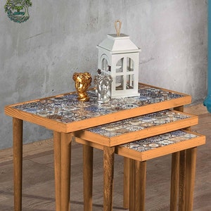 May include: A set of three nesting tables with wooden legs and tile-patterned tops. The tables are arranged in descending size, with the largest table in the front and the smallest table in the back. The tables are decorated with a variety of colorful tiles, including blue, green, and brown. The tables are sitting on a wooden floor.