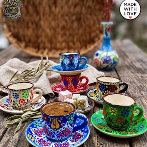 May include: A collection of ceramic Turkish coffee cups and saucers featuring vibrant, hand-painted floral patterns in blue, red, green, and black. The set includes multiple cups, some stacked, with one filled with dark coffee and small pieces of Turkish delight.