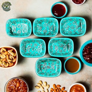 May include: A top-down shot showcases six turquoise ceramic dishes with white floral patterns. The dishes are rectangular with rounded corners. Small turquoise bowls hold sauces, and other bowls contain nuts and preserves. The arrangement is on a neutral surface.