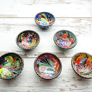 May include: Six small, colorful ceramic bowls with floral and bird designs. The bowls have a black rim and are arranged on a white wooden surface.
