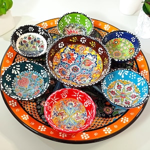 以下が含まれることがあります： 複雑な花柄が施されたカラフルなセラミックボウル8個セットが、オレンジと黒の丸いトレイに並べられています。ボウルは青、緑、赤、黄色の様々な色合いです。