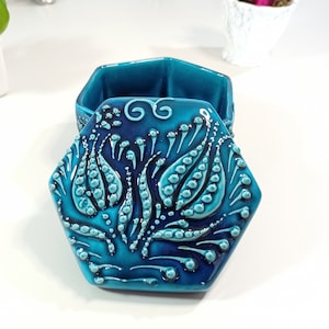 Peut inclure: Une boîte à bijoux hexagonale en céramique avec un glaçage bleu sarcelle et bleu foncé. Le couvercle présente un motif floral en relief avec un motif en spirale.
