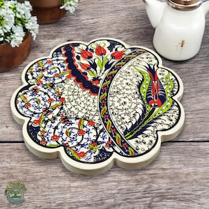 Puede incluir: Un posavasos decorativo de cerámica con borde festoneado. El posavasos presenta un vibrante diseño floral en rojo, azul y verde sobre un fondo blanco. El posavasos tiene aproximadamente 15 cm de diámetro.