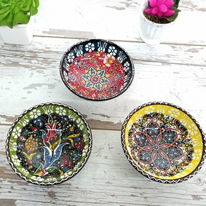 Puede incluir: Tres cuencos de cer&aacute;mica peque&ntilde;os y decorativos con intrincados dise&ntilde;os florales. Uno tiene un interior rojo con detalles en blanco y negro, otro tiene un dise&ntilde;o verde y negro, y el tercero tiene un interior amarillo. Los cuencos est&aacute;n sobre una superficie de madera blanca envejecida.