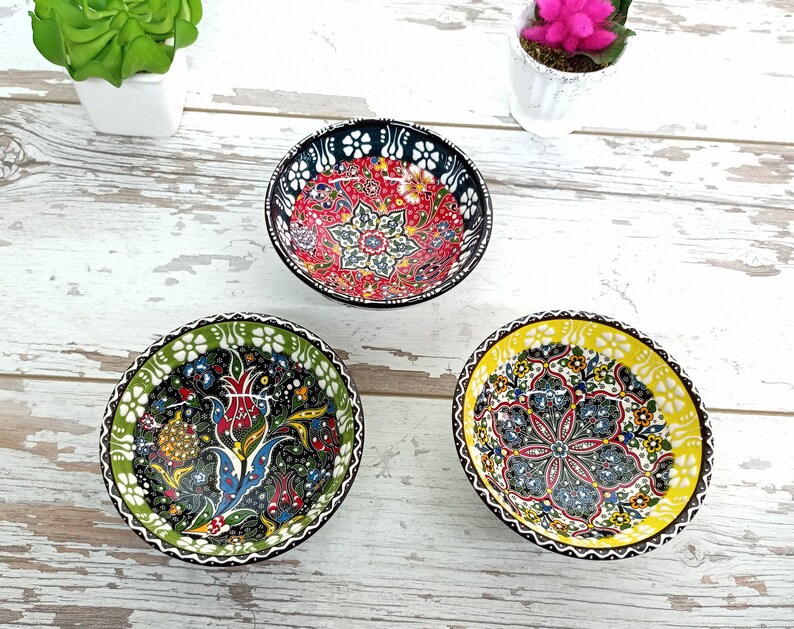 Puede incluir: Tres cuencos de cer&aacute;mica peque&ntilde;os y decorativos con intrincados dise&ntilde;os florales. Uno tiene un interior rojo con detalles en blanco y negro, otro tiene un dise&ntilde;o verde y negro, y el tercero tiene un interior amarillo. Los cuencos est&aacute;n sobre una superficie de madera blanca envejecida.