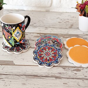 Puede incluir: Una taza con estampado floral en blanco y negro descansa sobre un posavasos a juego. Se muestran varios posavasos adicionales con coloridos dise&ntilde;os florales. Los posavasos tienen un borde festoneado blanco y una base naranja texturizada.
