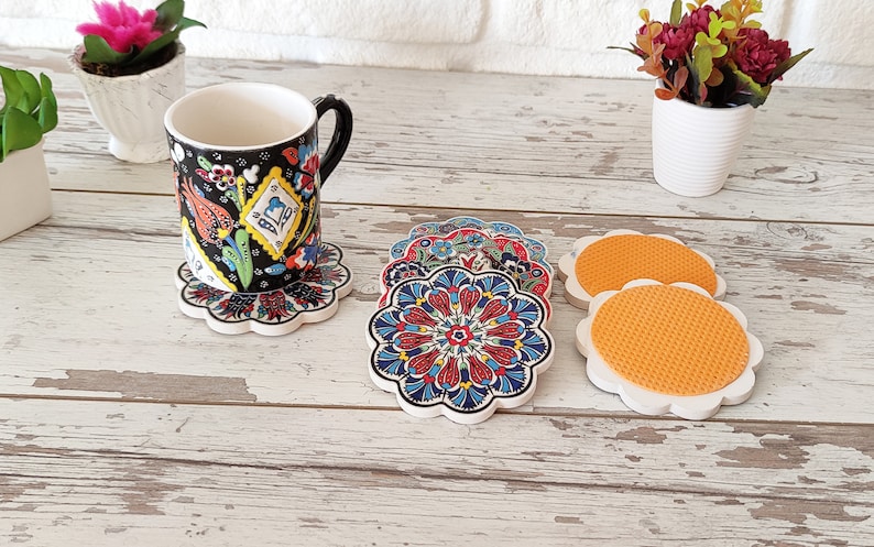 Puede incluir: Una taza con estampado floral en blanco y negro descansa sobre un posavasos a juego. Se muestran varios posavasos adicionales con coloridos dise&ntilde;os florales. Los posavasos tienen un borde festoneado blanco y una base naranja texturizada.