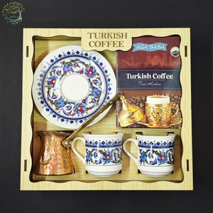 May include: A wooden gift box containing a Turkish coffee set. It includes a copper coffee pot, two white ceramic cups with blue and red floral patterns, a matching saucer, and a box of Aga Baba Turkish Coffee with a gold-toned cup illustration.
